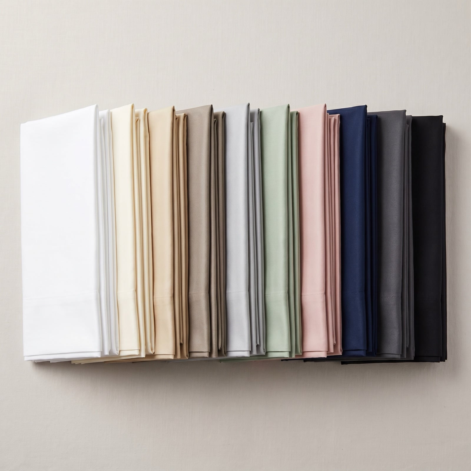 Ten folded Egyptian cotton sateen sheet sets fanned in a row showing the full color palette from pure white and ivory through sand, taupe, silver, sage, blush, navy, charcoal and black