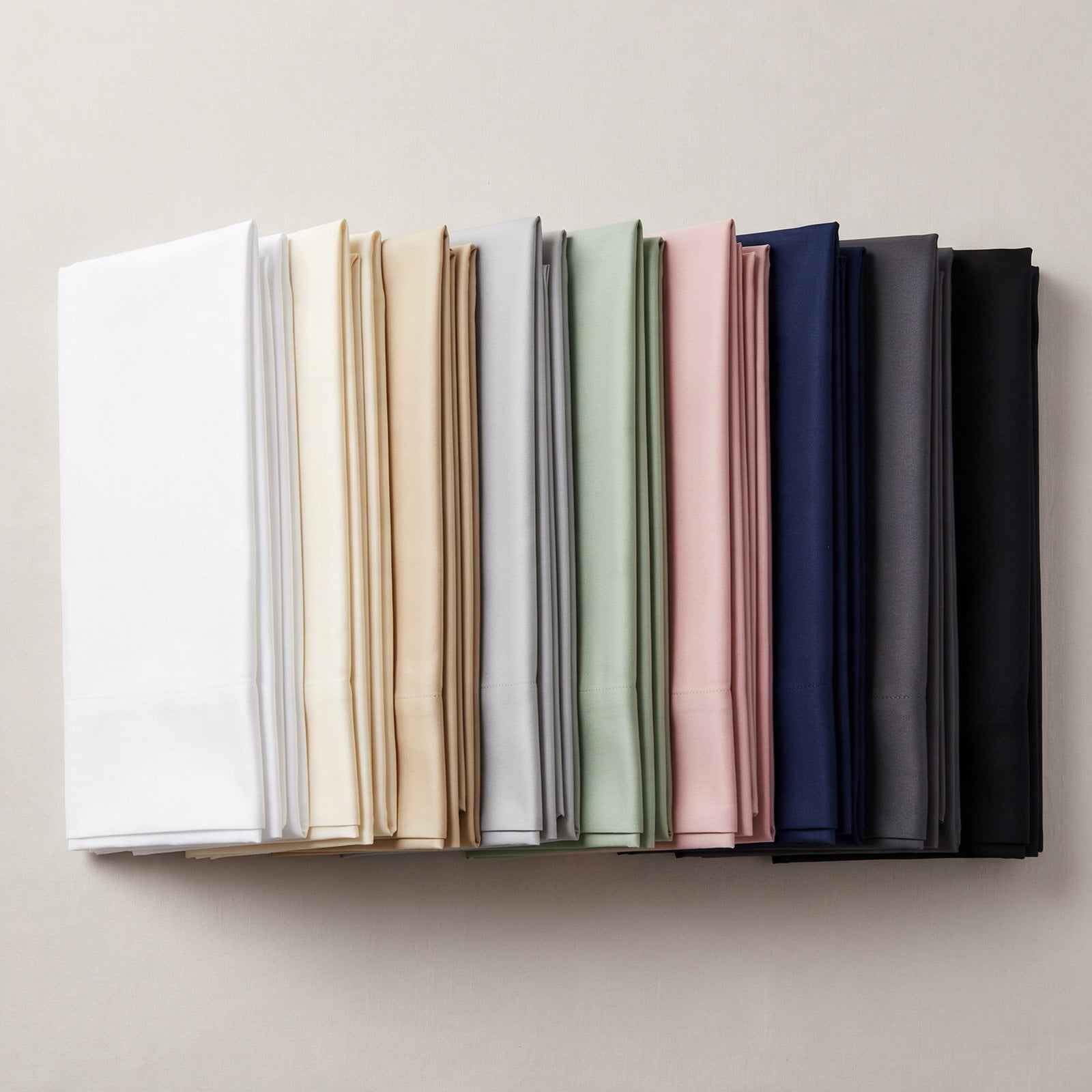 Folded Egyptian cotton sateen sheet sets fanned in a row showing the color palette from pure white and ivory through sand, silver, sage, blush, navy, charcoal and black